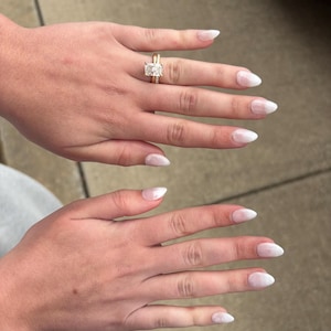 May include: A gold engagement ring with a large, rectangular diamond is shown on a finger. The ring is paired with a gold wedding band. The nails are painted with a white ombre design.