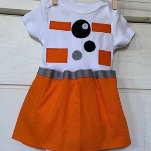May include: A white baby onesie with an orange and black design resembling a robot. The onesie has a grey ribbon around the waist and an orange skirt attached.