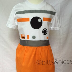 May include: A white t-shirt with an orange and gray BB-8 design. The shirt is paired with an orange skirt and a gray belt. The text "obitt&piece" is visible on the skirt.