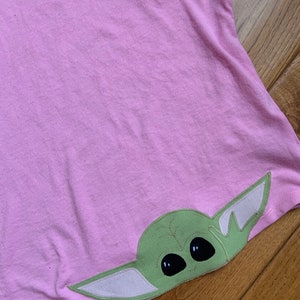 Pode incluir: Uma camiseta rosa com um personagem bordado verde e branco da franquia Star Wars. O personagem tem olhos grandes e orelhas pequenas.