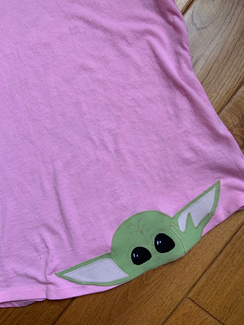Pode incluir: Uma camiseta rosa com um personagem bordado verde e branco da franquia Star Wars. O personagem tem olhos grandes e orelhas pequenas.