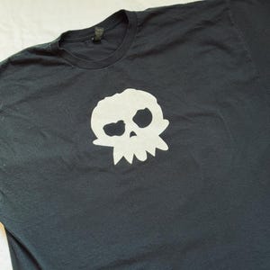 Puede incluir: Camiseta negra con un dise&ntilde;o de calavera blanca. El dise&ntilde;o de la calavera tiene cuencas oculares grandes y un borde inferior dentado. La camiseta est&aacute; hecha de un material suave y tiene cuello redondo.