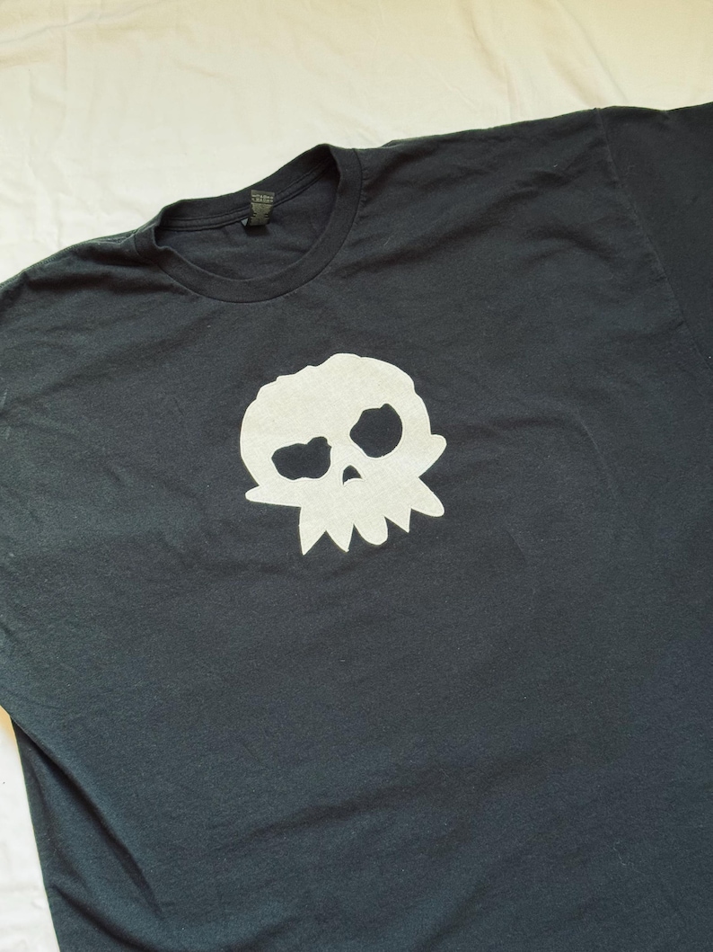 Puede incluir: Camiseta negra con un dise&ntilde;o de calavera blanca. El dise&ntilde;o de la calavera tiene cuencas oculares grandes y un borde inferior dentado. La camiseta est&aacute; hecha de un material suave y tiene cuello redondo.