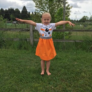 May include: A young girl wearing a white t-shirt with a black and white image of a robot on the front and an orange skirt. She is standing in a grassy field with a wooden fence in the background.