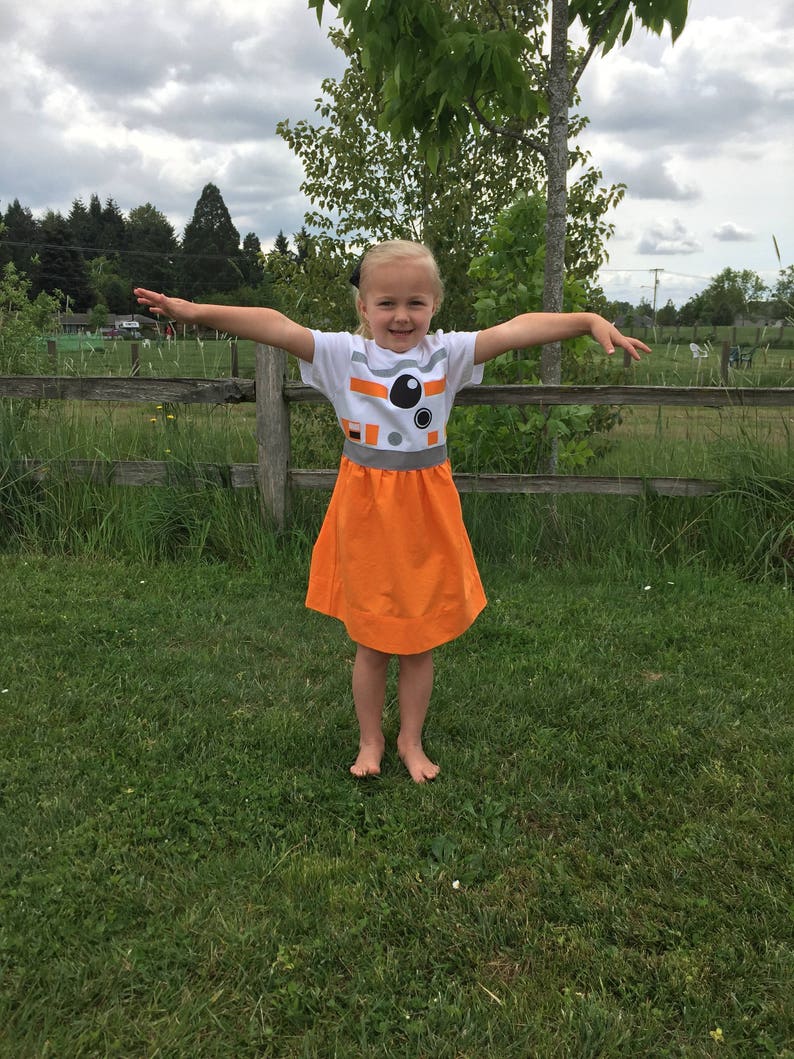 May include: A young girl wearing a white t-shirt with a black and white image of a robot on the front and an orange skirt. She is standing in a grassy field with a wooden fence in the background.