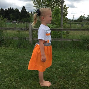 May include: A young girl wearing a white t-shirt with a grey and orange design and an orange skirt. The design on the shirt resembles the character BB-8 from Star Wars.