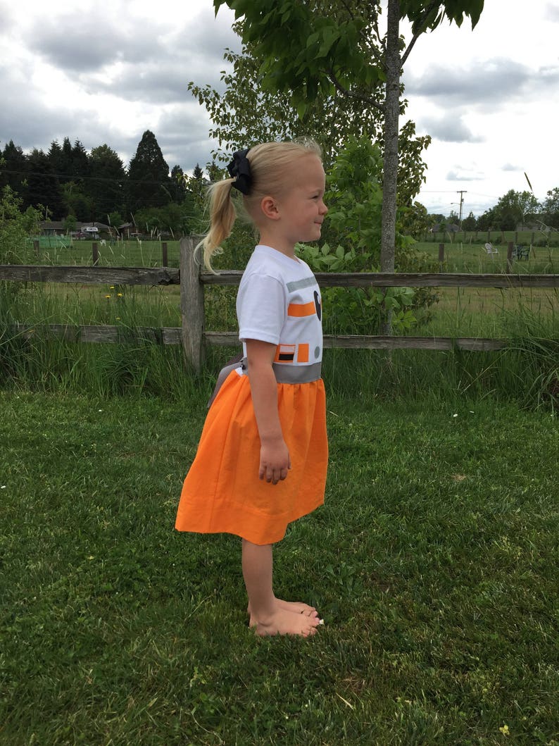 May include: A young girl wearing a white t-shirt with a grey and orange design and an orange skirt. The design on the shirt resembles the character BB-8 from Star Wars.