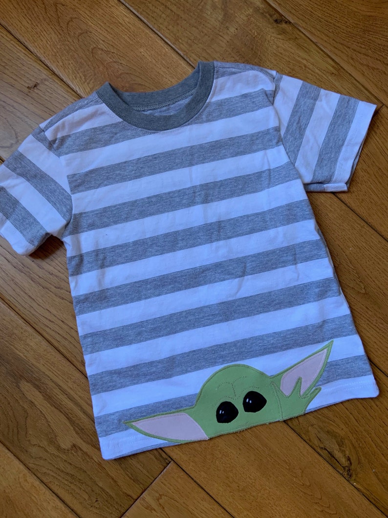 Pode incluir: Uma camiseta listrada cinza e branca com um aplique de personagem verde, rosa e preto. O personagem tem olhos grandes e uma cabe&ccedil;a pontiaguda.