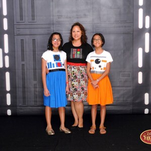 May include: Three people, two girls and a woman, are wearing Star Wars themed dresses. The girls are wearing blue and orange dresses with R2-D2 and BB-8 designs. The woman is wearing a black and white dress with a comic book pattern. The background is a black and white wall with a Star Wars theme.