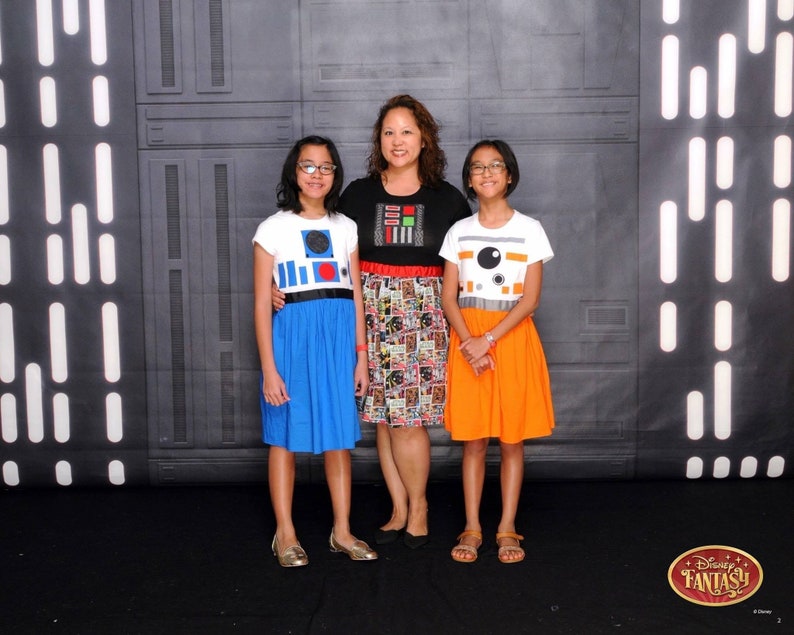 May include: Three people, two girls and a woman, are wearing Star Wars themed dresses. The girls are wearing blue and orange dresses with R2-D2 and BB-8 designs. The woman is wearing a black and white dress with a comic book pattern. The background is a black and white wall with a Star Wars theme.