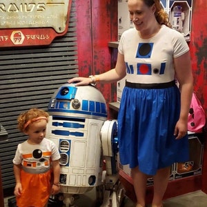 May include: A woman and a young girl stand in front of a white and blue R2-D2 robot. The woman is wearing a white shirt with a blue and red R2-D2 design and a blue skirt. The girl is wearing an orange and white dress with an R2-D2 design. The robot is standing on a gray floor. The sign behind them reads "Ramech Raid" in a stylized font.