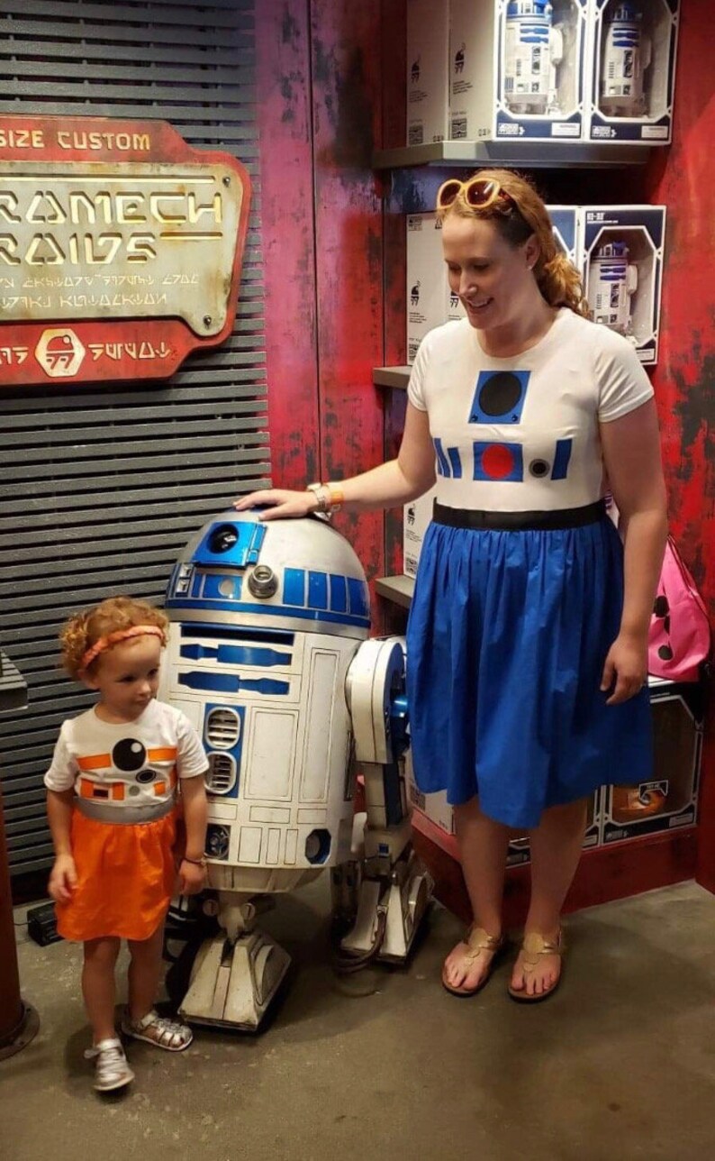 May include: A woman and a young girl stand in front of a white and blue R2-D2 robot. The woman is wearing a white shirt with a blue and red R2-D2 design and a blue skirt. The girl is wearing an orange and white dress with an R2-D2 design. The robot is standing on a gray floor. The sign behind them reads "Ramech Raid" in a stylized font.