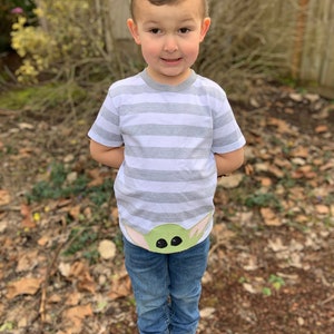 Pode incluir: Uma crian&ccedil;a pequena vestindo uma camiseta listrada cinza e branca com um personagem verde da franquia Star Wars na frente. A crian&ccedil;a tamb&eacute;m est&aacute; vestindo jeans azul e sapatos marrons.