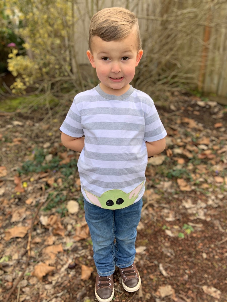 Pode incluir: Uma crian&ccedil;a pequena vestindo uma camiseta listrada cinza e branca com um personagem verde da franquia Star Wars na frente. A crian&ccedil;a tamb&eacute;m est&aacute; vestindo jeans azul e sapatos marrons.