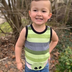 Pode incluir: Um menino pequeno vestindo uma camiseta sem mangas listrada de verde e cinza com um personagem de desenho animado na frente. O personagem tem olhos grandes e orelhas cor-de-rosa. O menino est&aacute; vestindo jeans azul.