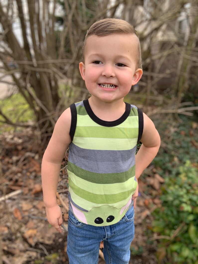 Pode incluir: Um menino pequeno vestindo uma camiseta sem mangas listrada de verde e cinza com um personagem de desenho animado na frente. O personagem tem olhos grandes e orelhas cor-de-rosa. O menino est&aacute; vestindo jeans azul.