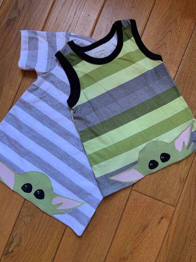 Pode incluir: Duas camisas listradas com um gr&aacute;fico de personagem verde. A camisa &agrave; esquerda &eacute; branca e cinza com mangas curtas. A camisa &agrave; direita &eacute; verde, cinza e branca sem mangas. Ambas as camisas t&ecirc;m um personagem verde com olhos grandes e orelhas cor-de-rosa.