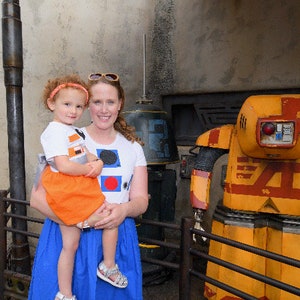 May include: A woman and a young girl are standing in front of a yellow and red robot. The robot has a large head with a red light and a black visor. The woman is wearing a white shirt with colorful geometric shapes and a blue skirt. The girl is wearing a white shirt with a black and orange design and an orange skirt.
