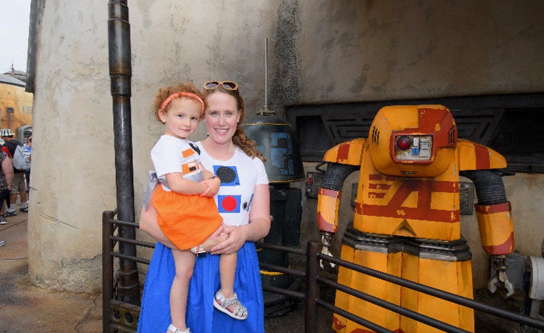 May include: A woman and a young girl are standing in front of a yellow and red robot. The robot has a large head with a red light and a black visor. The woman is wearing a white shirt with colorful geometric shapes and a blue skirt. The girl is wearing a white shirt with a black and orange design and an orange skirt.