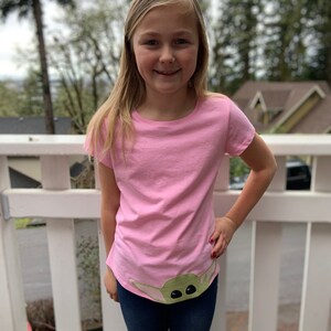 Pode incluir: Uma pessoa jovem vestindo uma camiseta rosa com um personagem verde da franquia Star Wars na parte inferior. O personagem tem olhos grandes e est&aacute; vestindo uma t&uacute;nica verde com capuz. A pessoa est&aacute; vestindo jeans azul e tem cabelo loiro comprido.