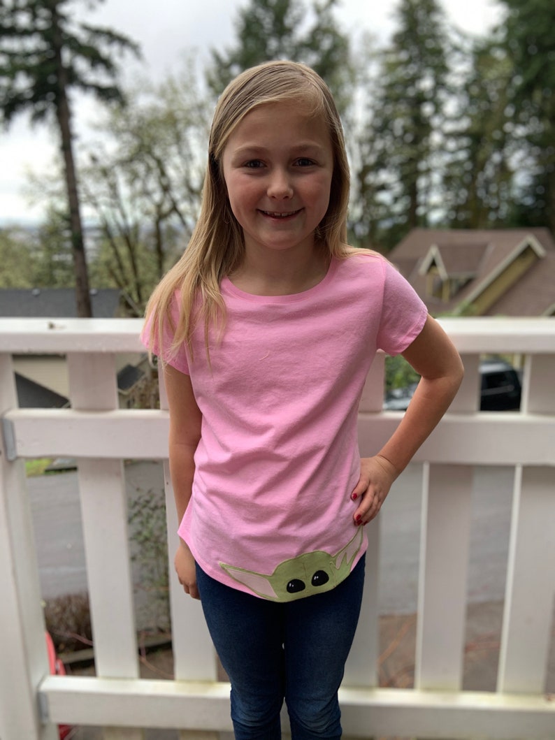 Pode incluir: Uma pessoa jovem vestindo uma camiseta rosa com um personagem verde da franquia Star Wars na parte inferior. O personagem tem olhos grandes e est&aacute; vestindo uma t&uacute;nica verde com capuz. A pessoa est&aacute; vestindo jeans azul e tem cabelo loiro comprido.