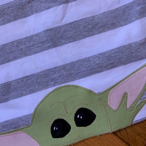 Pode incluir: Uma aplica&ccedil;&atilde;o de tecido verde e rosa de um personagem com olhos grandes, que se assemelha a um personagem fict&iacute;cio popular. A aplica&ccedil;&atilde;o &eacute; costurada em um fundo de tecido listrado.