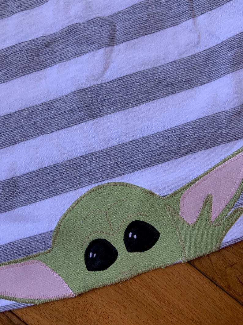 Pode incluir: Uma aplica&ccedil;&atilde;o de tecido verde e rosa de um personagem com olhos grandes, que se assemelha a um personagem fict&iacute;cio popular. A aplica&ccedil;&atilde;o &eacute; costurada em um fundo de tecido listrado.