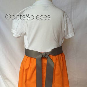 May include: A white t-shirt with a gray sash tied around the waist, worn over an orange skirt. The skirt is gathered at the waist and has a full, flowing shape.