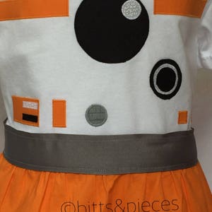 May include: A white dress with orange and gray accents, featuring an embroidered design of a robot character with a large black eye and a smaller black circle. The dress has a gray waistband and a full orange skirt. The text "&copy;bitts&pieces" is visible on the skirt.