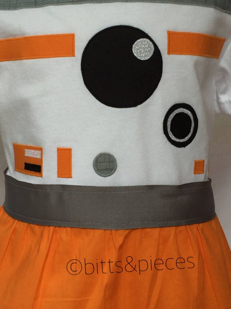May include: A white dress with orange and gray accents, featuring an embroidered design of a robot character with a large black eye and a smaller black circle. The dress has a gray waistband and a full orange skirt. The text "&copy;bitts&pieces" is visible on the skirt.
