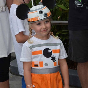 May include: A young girl wearing a white t-shirt with an orange and grey design of a robot. She is wearing a white and orange Mickey Mouse hat with black ears. The girl is wearing a matching orange skirt.