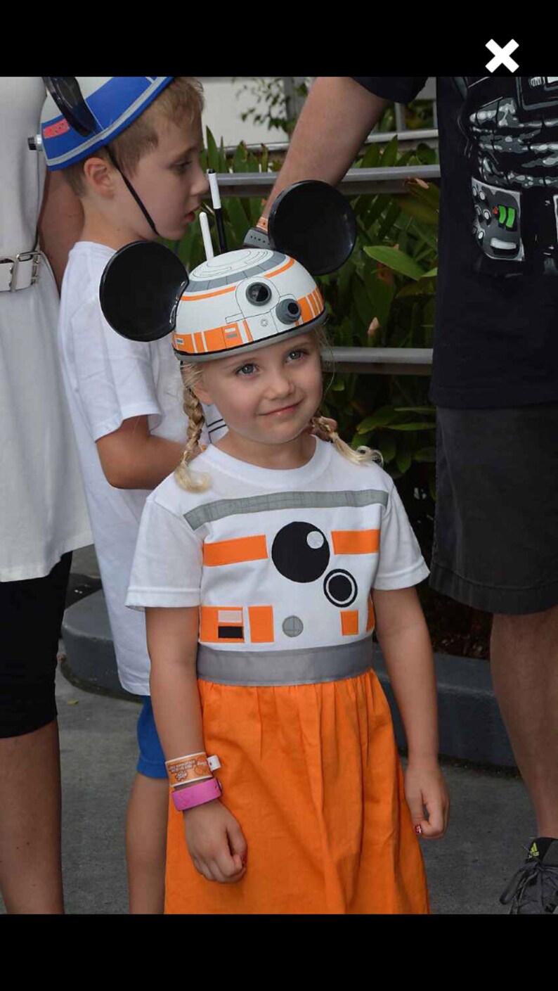 May include: A young girl wearing a white t-shirt with an orange and grey design of a robot. She is wearing a white and orange Mickey Mouse hat with black ears. The girl is wearing a matching orange skirt.
