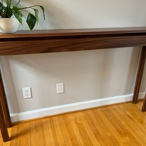 May include: A dark brown wooden console table with a hidden storage compartment. The table has a simple design with straight lines and a rectangular top. The table is standing on a hardwood floor.