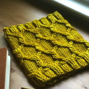 May include: Mustard yellow knitted cowl with a textured diamond pattern. The chunky knit scarf is made of thick yarn and is shown on a wooden surface with knitting needles and a notebook.