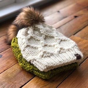 Op de afbeelding: Een witte gebreide beanie met een bruine nep bont pompon. De beanie is omgeslagen en heeft een groene gebreide band eronder.