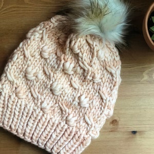 May include: A knitted beanie hat with a fluffy pom pom in a light peach color.