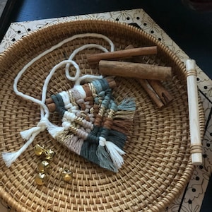 May include: A woven macrame wall hanging with a geometric design in shades of brown, beige, white, and green. The hanging has a fringe of white yarn and is displayed on a woven tray with cinnamon sticks and gold bells.
