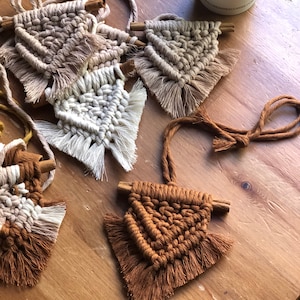 Op de afbeelding: Vier macramé wandhangers in verschillende tinten bruin, beige en wit. Elke hanger heeft een driehoekige vorm met franjes en een kaneelstokje dat aan de bovenkant is bevestigd.