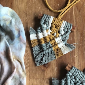 May include: Two macrame wall hangings with fringe, one in grey, white, and mustard yellow, and the other in grey. The grey, white, and mustard yellow wall hanging is attached to a cord.