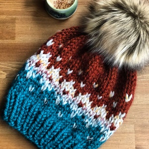 May include: A knitted hat with a faux fur pom-pom. The hat has a colour block design with teal, rust, and white sections. A mug of coffee sits in the background on a wooden surface.