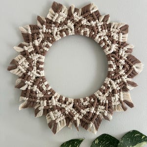 Op de afbeelding: Een bruine en crèmekleurige macramé wandhanger in de vorm van een zon met een cirkelvormig midden. De macramé is gemaakt met een dik koord en heeft een franje aan de randen.