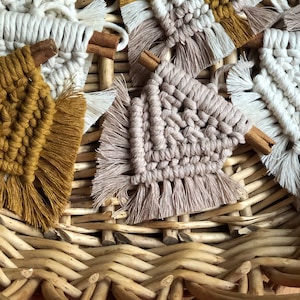 May include: A collection of small, handmade macrame wall hangings with fringe, each featuring a different colour combination of beige, white, and mustard yellow. The hangings are displayed in a wicker basket.