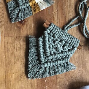 May include: A light blue macrame wall hanging with a fringe border. The hanging is made with thick rope and has a wooden dowel at the top.