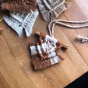 Puede incluir: Primer plano de varios posavasos de macramé hechos a mano en tonos marrón y crema. Cada posavasos presenta un diseño geométrico único con bordes con flecos y un acento de palito de canela. Los posavasos están dispuestos sobre una superficie de madera.