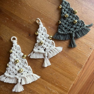 May include: Three handmade macrame Christmas tree decorations with gold bells. The trees are in white, green, and natural colours.