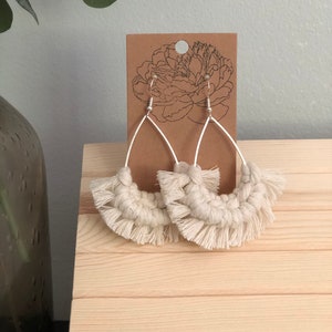 May include: A pair of cream-colored macrame earrings with fringe details hanging from a brown card with a floral design.