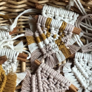 Może przedstawiać: Zbliżenie na kilka ręcznie robionych makramowych wiszących ozdób ściennych z frędzlami. Ozdoby wykonane są z białej, beżowej i musztardowej przędzy. Każda ozdoba ozdobiona jest patyczkiem cynamonu.