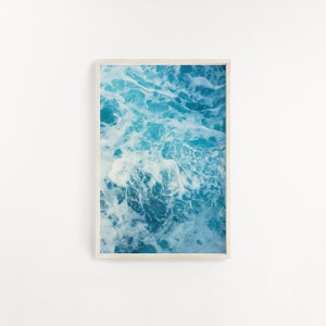 May include: A framed print featuring an abstract image of ocean water. The artwork showcases a close-up view of churning turquoise and white waves, creating a dynamic and textured visual effect. The print is encased in a light-colored wooden frame.