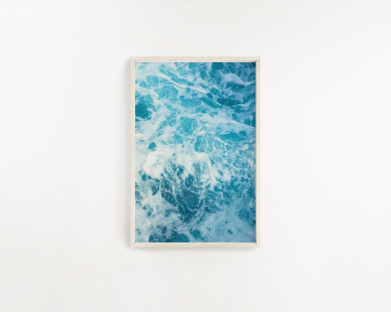 May include: A framed print featuring an abstract image of ocean water. The artwork showcases a close-up view of churning turquoise and white waves, creating a dynamic and textured visual effect. The print is encased in a light-colored wooden frame.