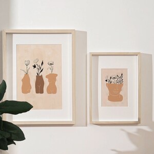 Pu&ograve; includere: Due stampe incorniciate con disegni minimalisti di figure stilizzate con fiori che crescono dalle loro teste e corpi. Le stampe sono su una parete bianca con una pianta verde in primo piano.